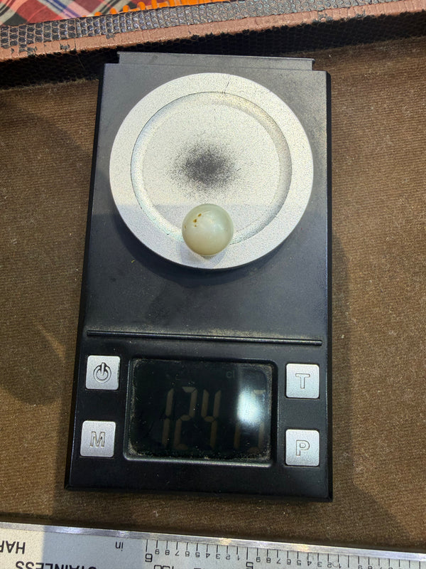 A small round pale green bead or gemstone moti placed on a digital pocket scale displaying 124 grams, set against a brown textured background.